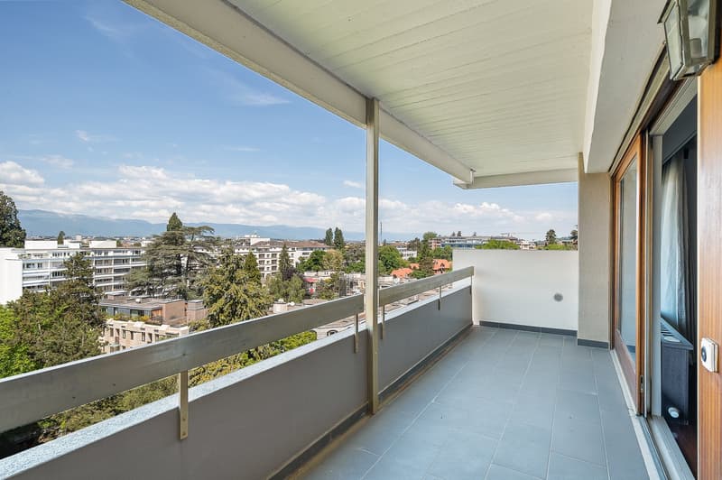 Spacious balcony with a panoramic view of the city, surrounded by trees and buildings. The balcony has a tiled floor and a glass railing. The apartment appears to be on a high floor, providing an elevated perspective of the urban landscape.