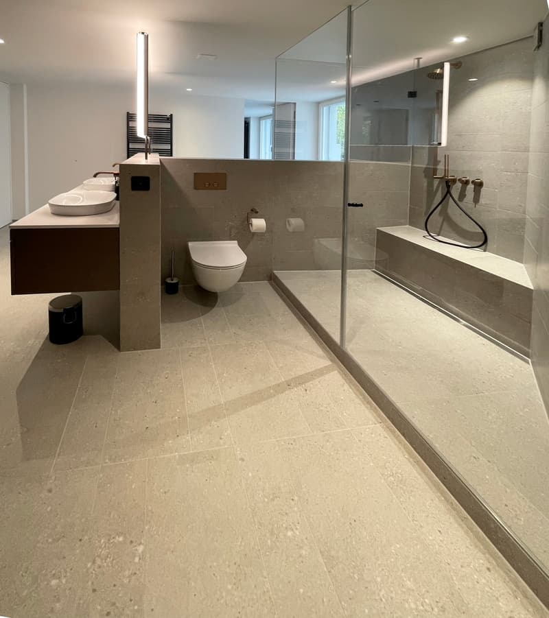 Large bathroom with modern design, beige tile flooring, vanity with double sinks, toilet, and glass shower enclosure