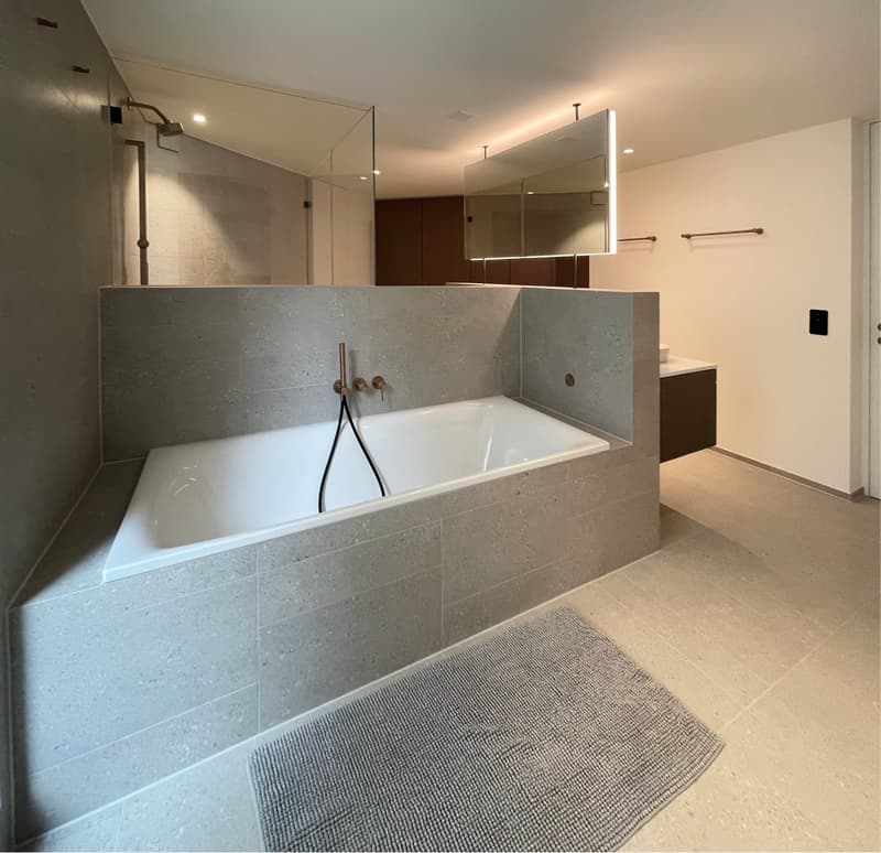 Large bathroom with a deep soaking tub, gray tile walls, built-in vanity with mirror, and modern fixtures