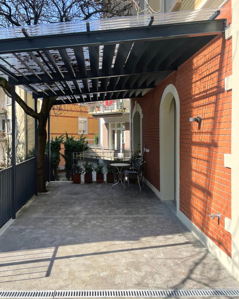 Two-story brick building with a covered patio area, featuring a metal pergola structure and a balcony above. The patio has a concrete floor and outdoor furniture, and the building has arched windows and an entryway.