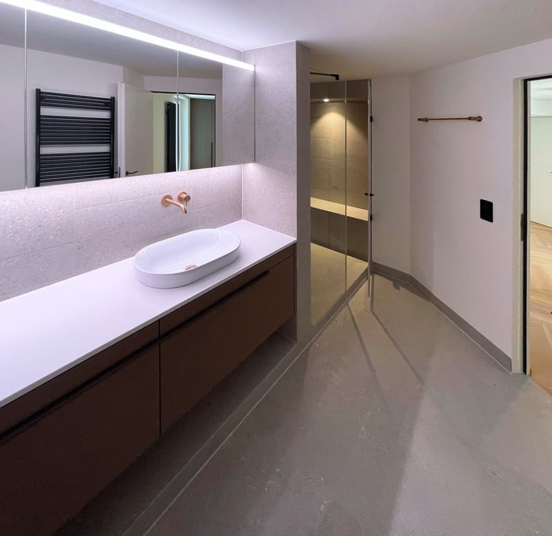 The image shows a modern and spacious bathroom with a minimalist design. The room features a large vanity with a white countertop and a vessel sink. The vanity has dark brown drawers, providing ample storage space. The bathroom also has a large mirror abo