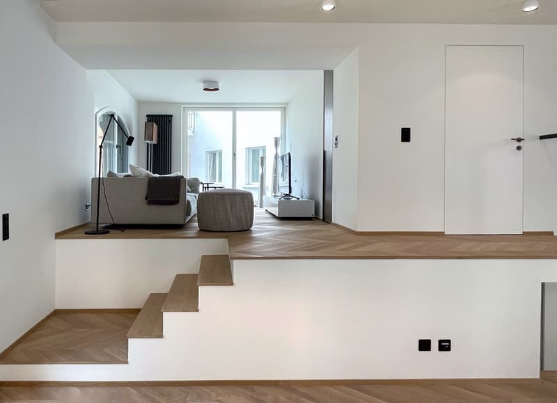 Spacious open-plan living area with raised platform, hardwood flooring, large windows, gray sofa, and minimalist decor