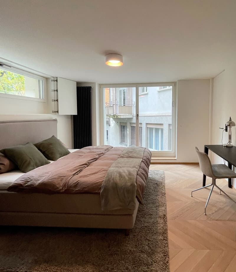 Spacious bedroom with large windows, hardwood flooring, a plush bed with brown and green bedding, and a desk with a chair near the window.