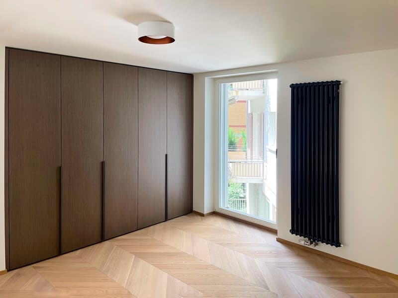 Large dark wood wardrobe, hardwood herringbone floor, large window with black curtains, radiator