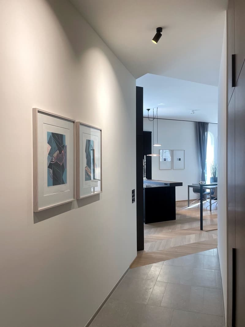 The image shows a hallway with white walls, a tiled floor, and framed artwork on the walls. There is a black light fixture on the ceiling and a view of a room with a table and chairs in the background.