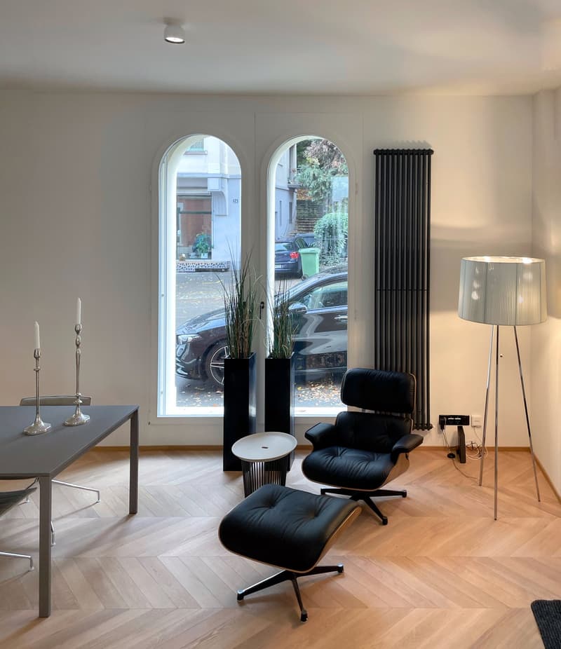 Spacious living room with arched windows, hardwood floors, black leather lounge chair, floor lamp, and a metal desk with decor items.