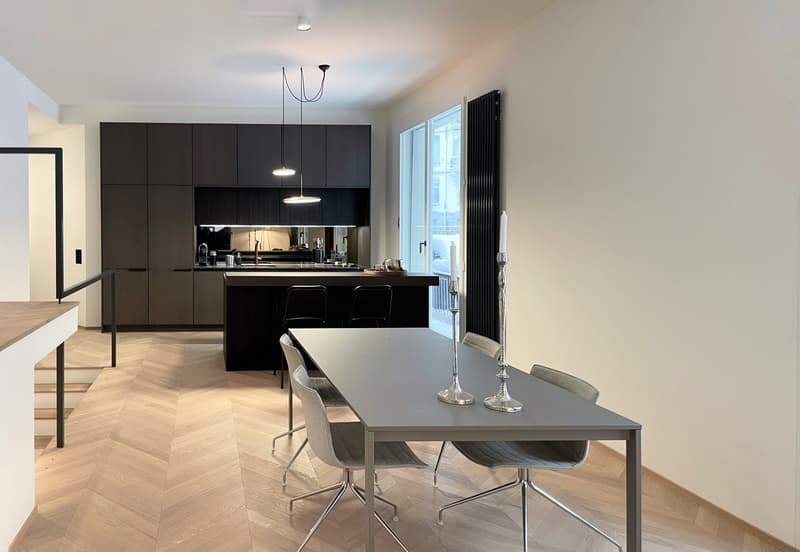 Modern open-concept kitchen and dining area with dark cabinets, stainless steel appliances, a large island with bar stools, and a dining table with chairs. The room has hardwood floors and large windows providing natural light.