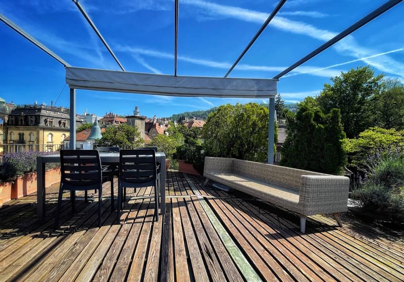 Wooden deck with outdoor furniture, including a sofa and chairs, overlooking a scenic city view with trees and buildings in the background. The terrace is covered by a retractable canopy or awning.