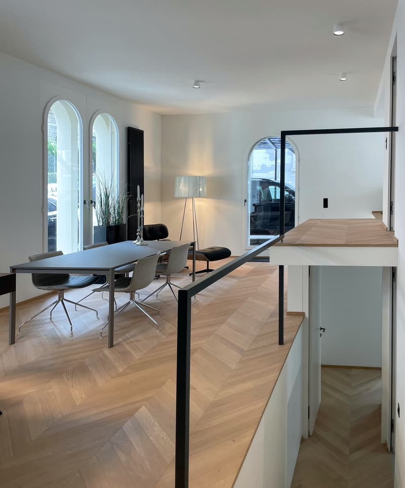 The image shows a modern and spacious dining room with a large dining table and chairs. The room has arched windows that provide natural light and a view of the exterior. The floor is made of light-colored wood in a herringbone pattern, adding a stylish t