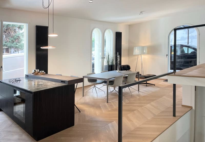 Spacious open-plan living and dining area with hardwood flooring, large windows, black radiators, modern pendant lighting, dining table with chairs, and a kitchen island with a built-in dishwasher.