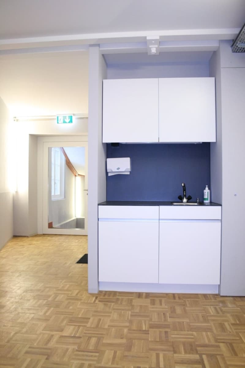 Compact kitchen with white cabinets, dark blue backsplash, and a dishwasher visible. Hardwood flooring and an open doorway leading to another room.