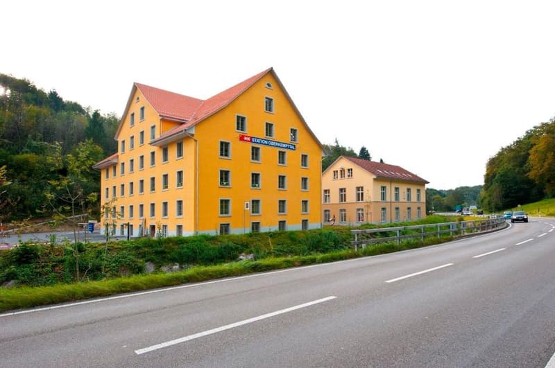 Two-story yellow building, red roof, multiple windows, sign reading 'Station Oberkempttal', located on a road with cars passing by, surrounded by greenery and trees.