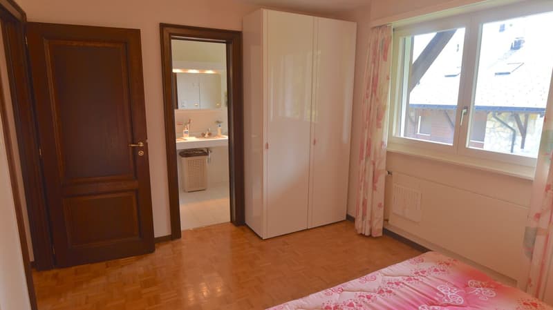 The image shows a bedroom with a wooden door, a large wardrobe, and a window. The room has a hardwood floor and the walls are painted in a light color. There is a bed with a floral patterned bedspread visible in the room.