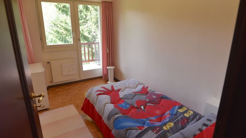 Bedroom with hardwood floor, white walls, large windows overlooking a balcony, and a bed with a colorful patterned comforter.