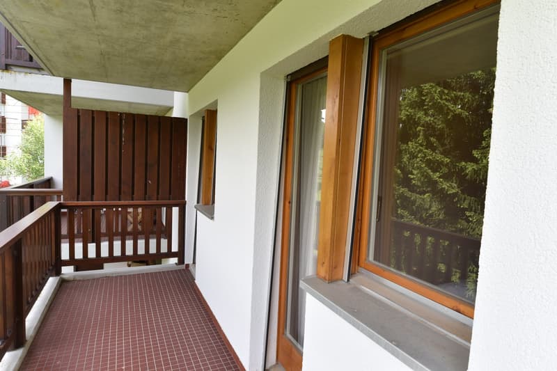 Covered balcony with wooden railings, concrete ceiling, and large windows overlooking a forested area