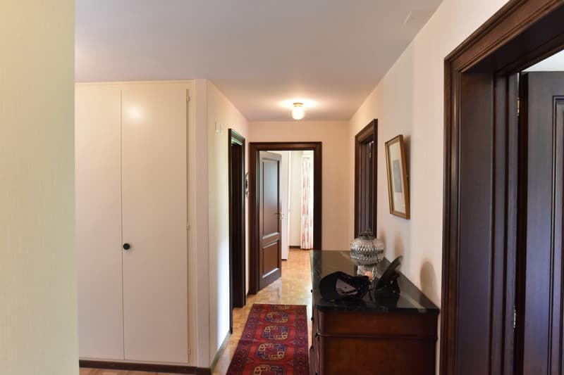 The image shows a hallway with a wooden floor, white walls, and a wooden door frame. There is a wooden cabinet or dresser against the wall, with a decorative vase on top. The hallway leads to other rooms, with additional doors visible.
