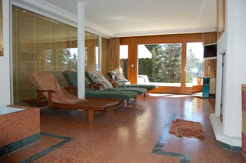 Spacious living room with large windows overlooking a forested area, wooden floors, comfortable lounge chairs, and a sliding glass door leading to a balcony or terrace.