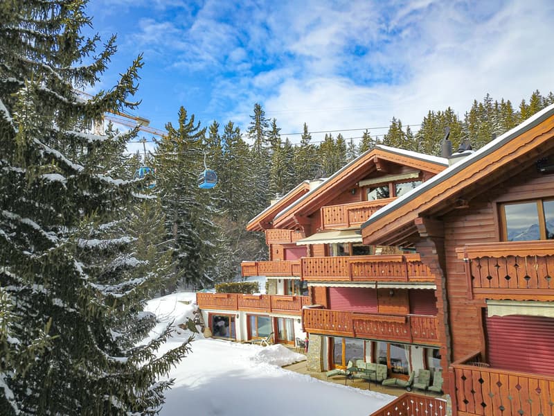 A snowy mountain scene with a ski resort featuring wooden chalets with balconies, surrounded by pine trees and a ski lift in the background.