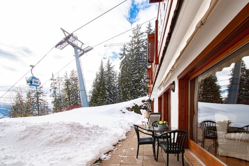 The image shows a ski resort with a ski lift in the background and a snow-covered landscape. The building in the foreground has a balcony or terrace with outdoor seating, suggesting it is a hotel or lodge at the ski resort.