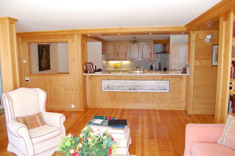 Spacious living room with wooden walls and floors, a fireplace, a white armchair, and a coffee table with books and flowers. The room also has a kitchen area with wooden cabinets and a countertop, as well as a framed artwork on the wall.