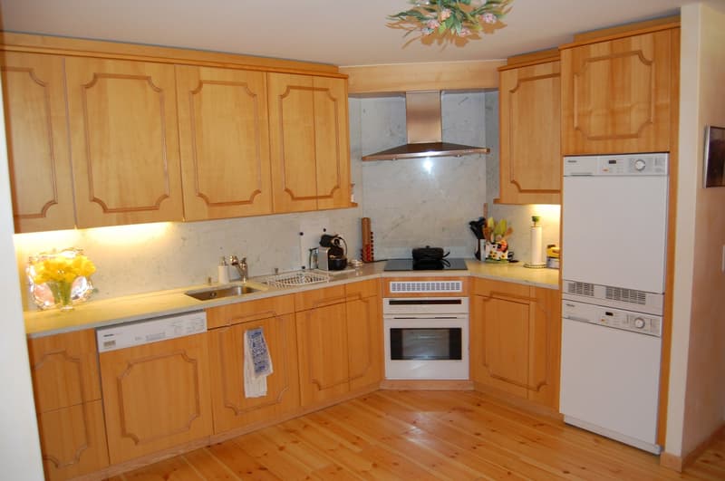 Spacious kitchen with wooden cabinets, marble countertops, stainless steel appliances including a dishwasher and washing machine, and hardwood flooring.
