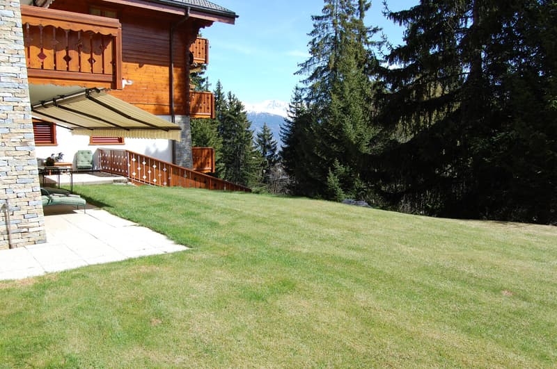 2-story wooden chalet-style house with stone exterior, large balcony, raised ground floor, surrounded by lush green lawn and pine trees, with a view of mountains in the background