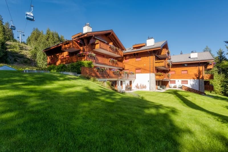 Multi-story wooden chalet-style building with a red roof, surrounded by lush green grass and trees, with a chairlift visible in the background.