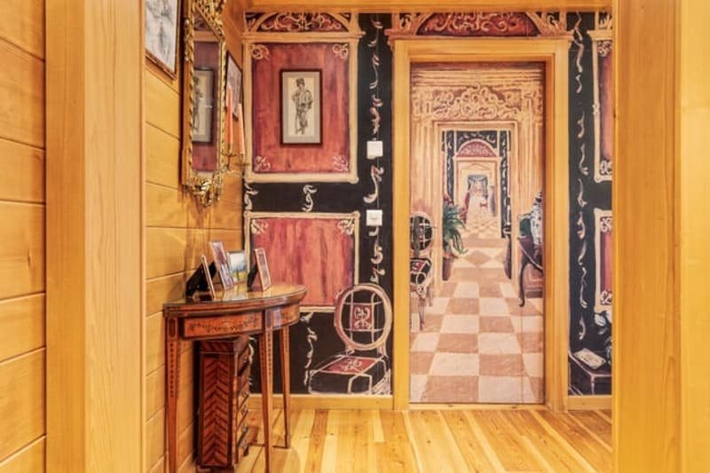 Ornate and decorative interior with wooden walls, checkered floor tiles, antique furniture, and framed artwork on the walls. The room appears to lead into another room or hallway through an ornate doorway.