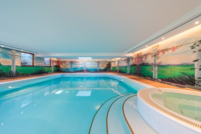 Large indoor swimming pool with tiled walls and floors, surrounded by glass windows providing a scenic view, and a hot tub or jacuzzi area.
