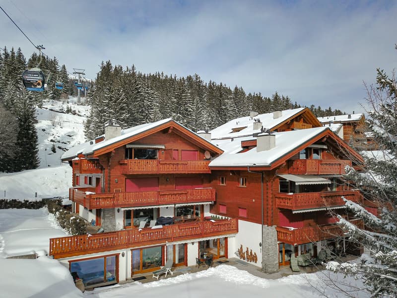 A group of wooden chalets with red roofs and balconies, surrounded by snow-covered pine trees and a ski lift in the background. The chalets appear to be part of a ski resort located in a mountainous, snowy environment.