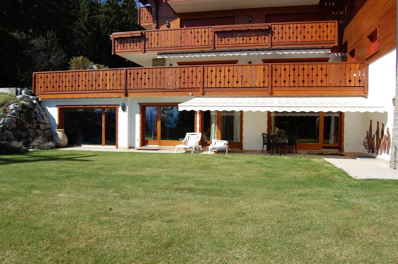 2-story wooden chalet-style house with a balcony, garage, and parking space. The house has large windows and a covered patio area with outdoor furniture. The property is surrounded by a well-maintained lawn and landscaping.