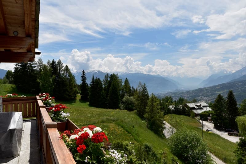 The image shows a scenic mountain landscape with a wooden chalet or cabin in the foreground. The cabin has a balcony or terrace overlooking the lush green hills and forests, with snow-capped mountains visible in the distance. The sky is filled with fluffy