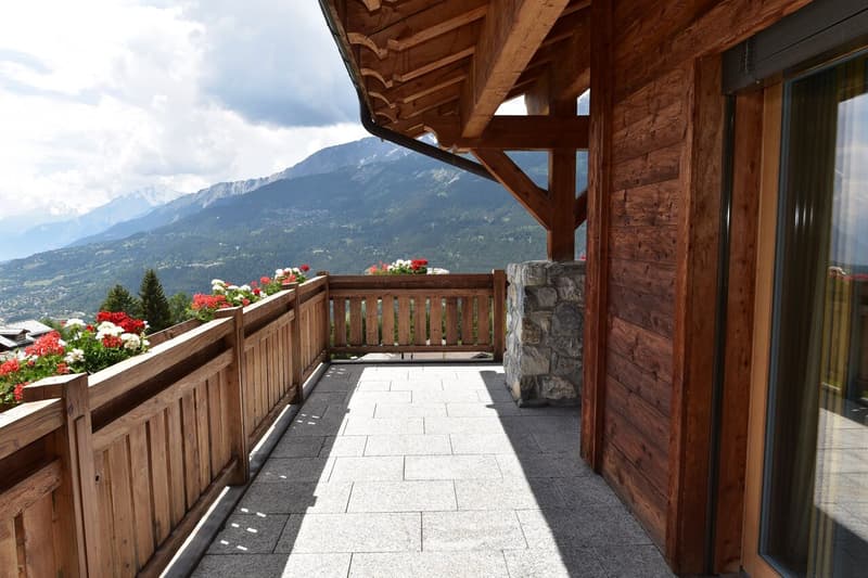 Wooden balcony with stone flooring, overlooking a mountainous landscape with pine trees. The balcony has a wooden railing and is decorated with potted flowers.