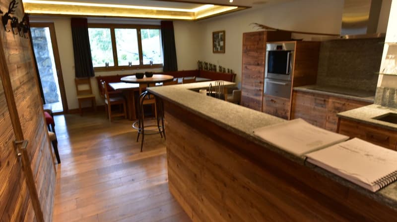 Large open-concept kitchen and dining area with wooden walls and floors, a stone fireplace, a dining table with chairs, a kitchen counter with bar stools, and built-in wooden cabinetry and appliances including a dishwasher.
