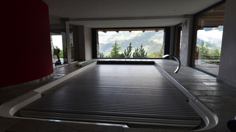 Large indoor pool with a panoramic view of the mountains and pine trees through the large windows. The pool has a sleek, modern design with a black tiled floor and a curved shape.