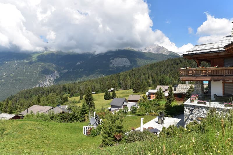 The image shows a scenic mountain landscape with a wooden chalet-style building in the foreground. The building has a balcony or terrace overlooking the lush green valley below, with a backdrop of snow-capped mountains and cloudy skies. The surrounding ar