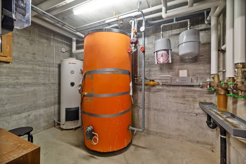 The image shows a utility room with an orange boiler or water heater, along with other plumbing equipment and a water heater on the side.