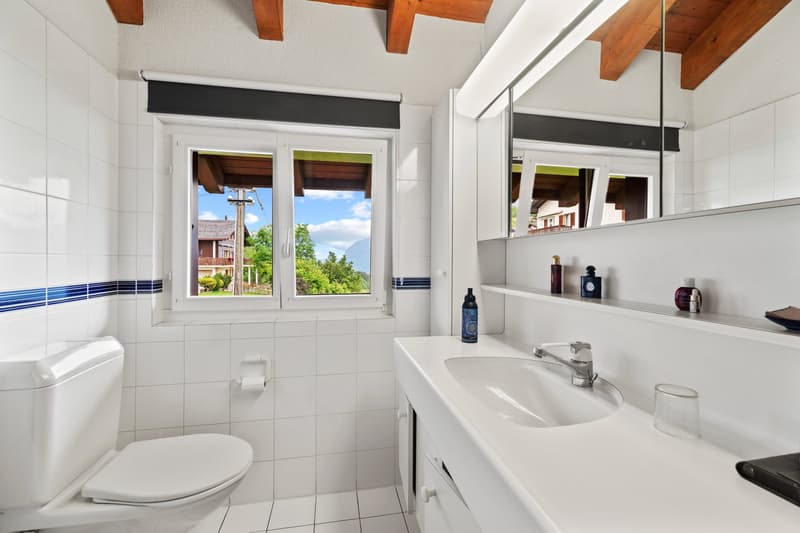 Spacious bathroom with white tiles, wooden ceiling beams, large window with scenic view, toilet, sink, and vanity mirror