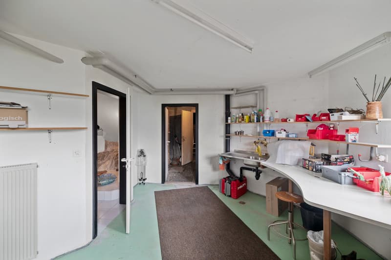 The image shows a garage or workshop space with various tools, equipment, and storage shelves. The room has a concrete floor and white walls, with a doorway leading to another room. There are several red containers and other items stored on the shelves.