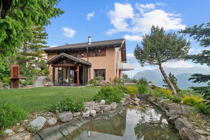 2-story house with wooden and glass architecture, surrounded by lush greenery, a small pond, and a scenic mountain view in the background