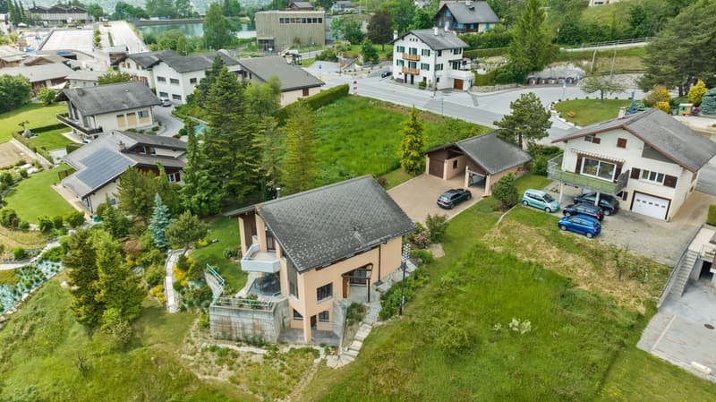 This image shows a residential area with a mix of houses, apartments, and other buildings. The area is surrounded by lush greenery, including trees and well-maintained lawns. There are several cars parked in the area, indicating the presence of parking sp