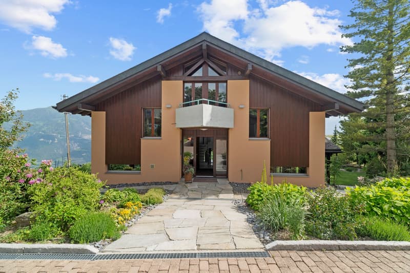 2-story wooden house with a slanted roof, large windows, and a balcony. The exterior is a combination of wood and stucco. The house is surrounded by a well-maintained garden with a stone pathway leading to the entrance.