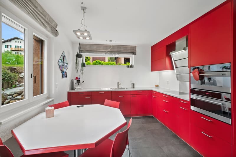 Modern kitchen with red cabinets, white countertops, large windows, dining table, and appliances including a dishwasher