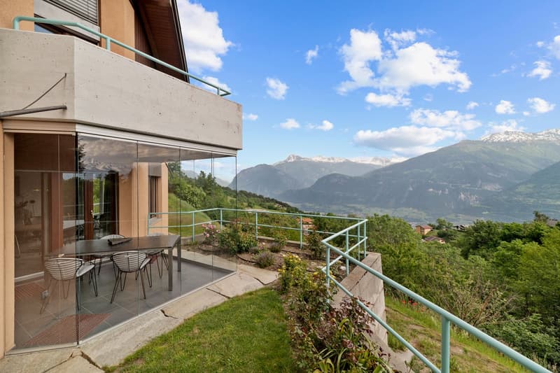 2-story modern house with a balcony/terrace overlooking a scenic mountain landscape. The house has large glass windows and doors that provide panoramic views of the surrounding nature. The balcony has a dining area with a table and chairs. The exterior fe
