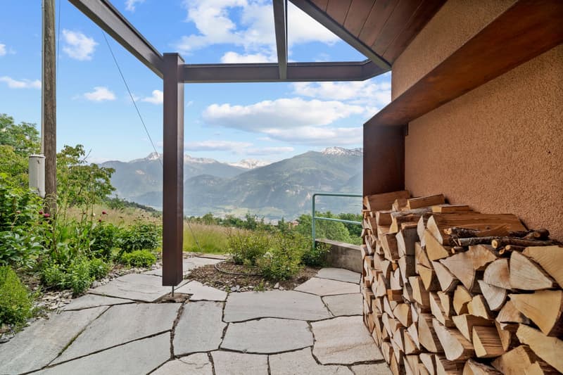 The image shows an outdoor patio or deck area with a stunning mountain view in the background. The patio has a wooden roof structure and a stone floor. There is a stack of firewood visible on the side. The surrounding landscape includes lush greenery and
