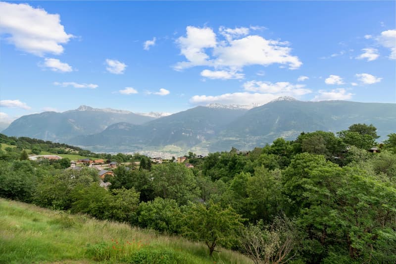 The image shows a scenic mountain landscape with a village nestled among the lush green trees and vegetation. The snow-capped peaks of the mountains can be seen in the distance under a blue sky with fluffy white clouds. The scene appears to be in a rural,