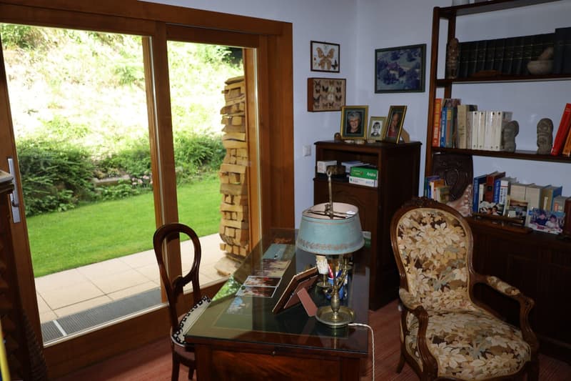 Large windows overlooking a garden, wooden furniture including a desk and armchair, shelves with books and decorative items, framed artwork on the walls
