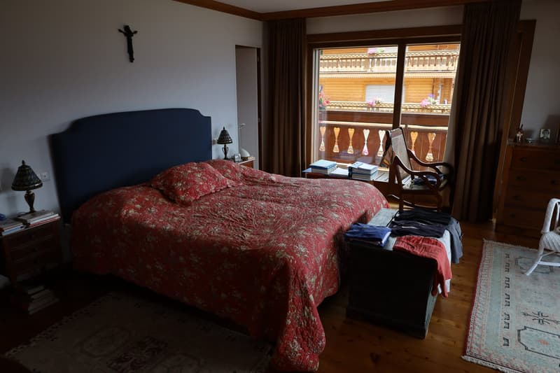 Large bedroom with a king-size bed, wooden furniture, and a balcony overlooking a scenic view. The room has a cozy and traditional decor with a crucifix on the wall.