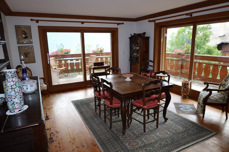 Spacious dining area with wooden furniture, including a large dining table and chairs with red cushions. The room has large windows overlooking a balcony or terrace with views of greenery outside. The walls feature wooden trim, and there is a cabinet or a