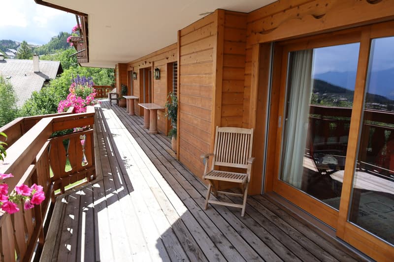 Wooden balcony with railing, wooden chairs, potted plants, and large windows overlooking a scenic mountain landscape